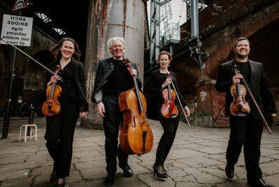 Members of the Dante Quartet hold their instruments in an industrial area of a city and smile widely.